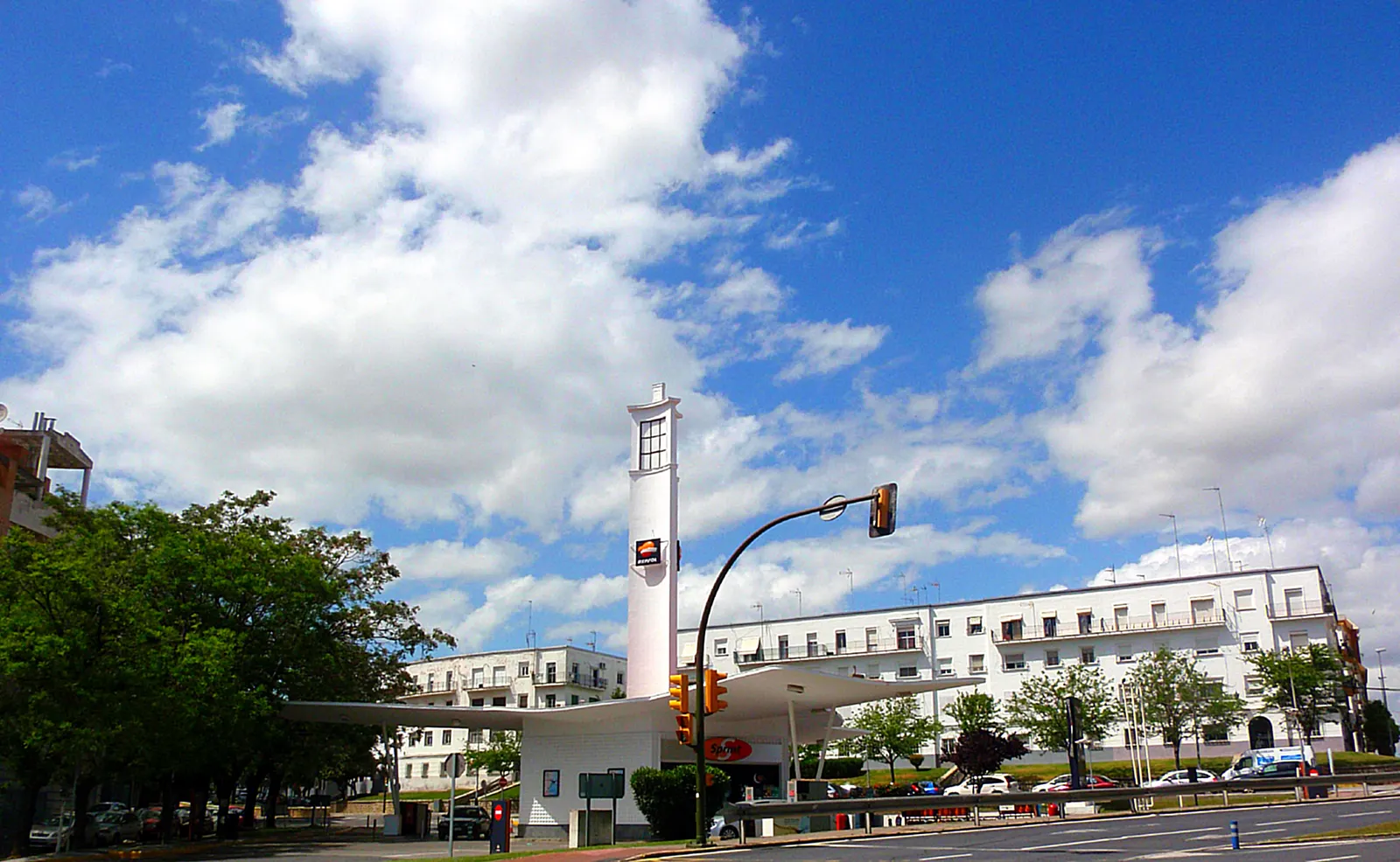 Estación de servicio con marquesina blanca y una torre-mástil rectangular con el logotipo de Repsol, junto a una avenida con coches y bloques de viviendas al fondo