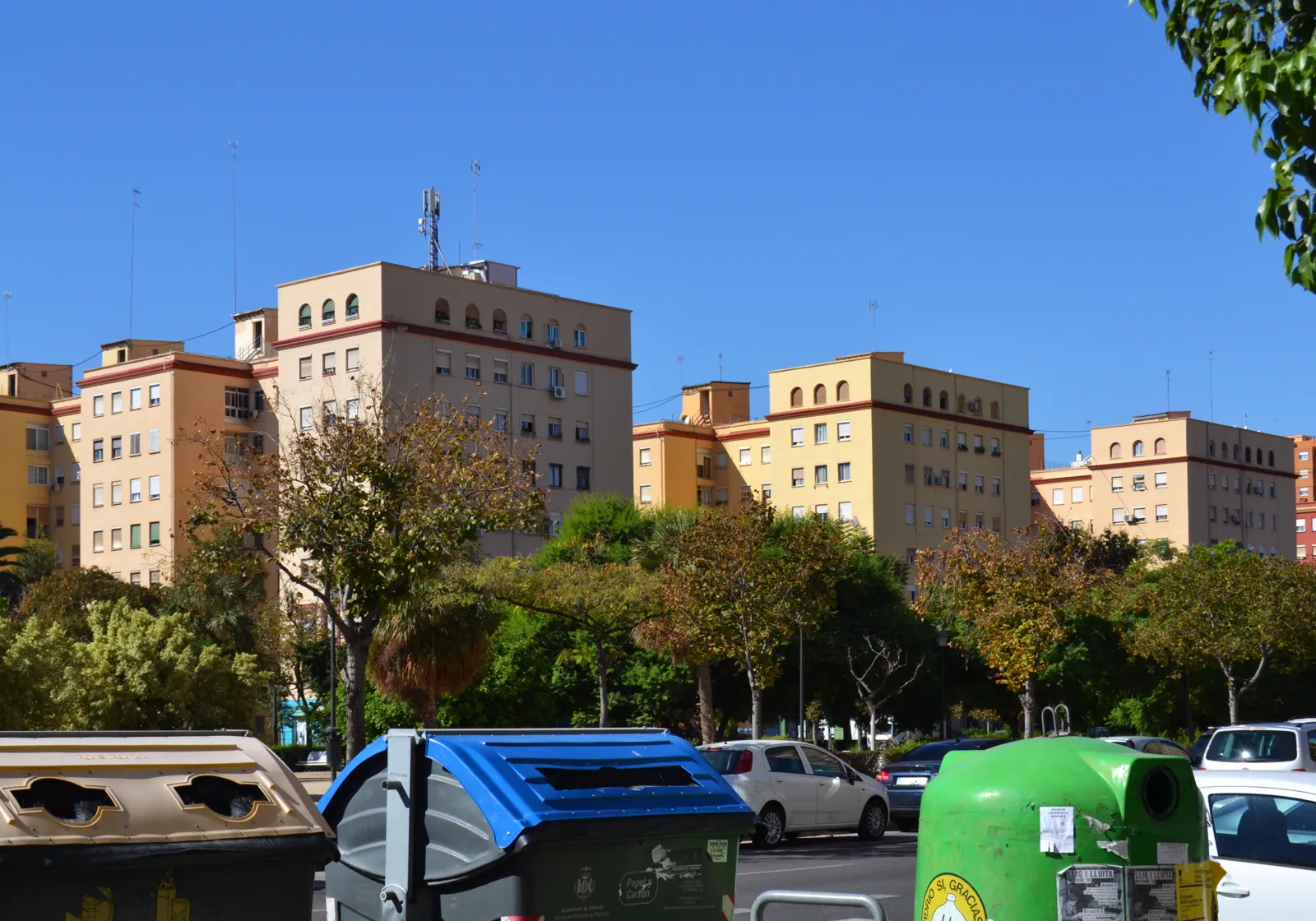 Hilera de bloques de viviendas de ladrillo de unas diez alturas, con balcones y toldos, vistos desde una calle ancha con coches aparcados y árboles jóvenes en la acera.