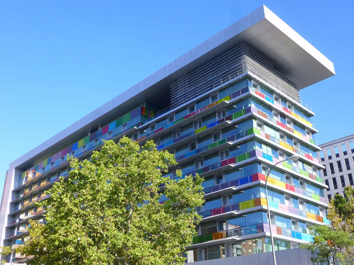 Edificio de oficinas de una decena de plantas fotografiado desde la acera, con la fachada cubierta de paneles de vidrio de colores —azules, verdes, rojos y amarillos— y una gran visera blanca volada sobre la azotea, con un árbol en primer plano y cielo despejado.