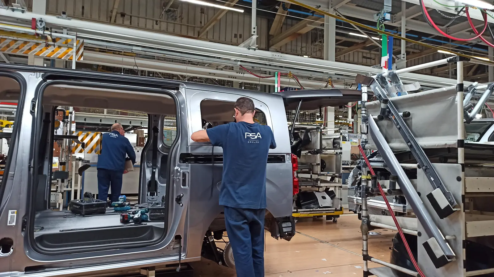 Dos operarios con polo azul y el logotipo PSA trabajan en la cadena de montaje de una fábrica de automóviles, junto a carrocerías grises de furgoneta con las puertas laterales abiertas.