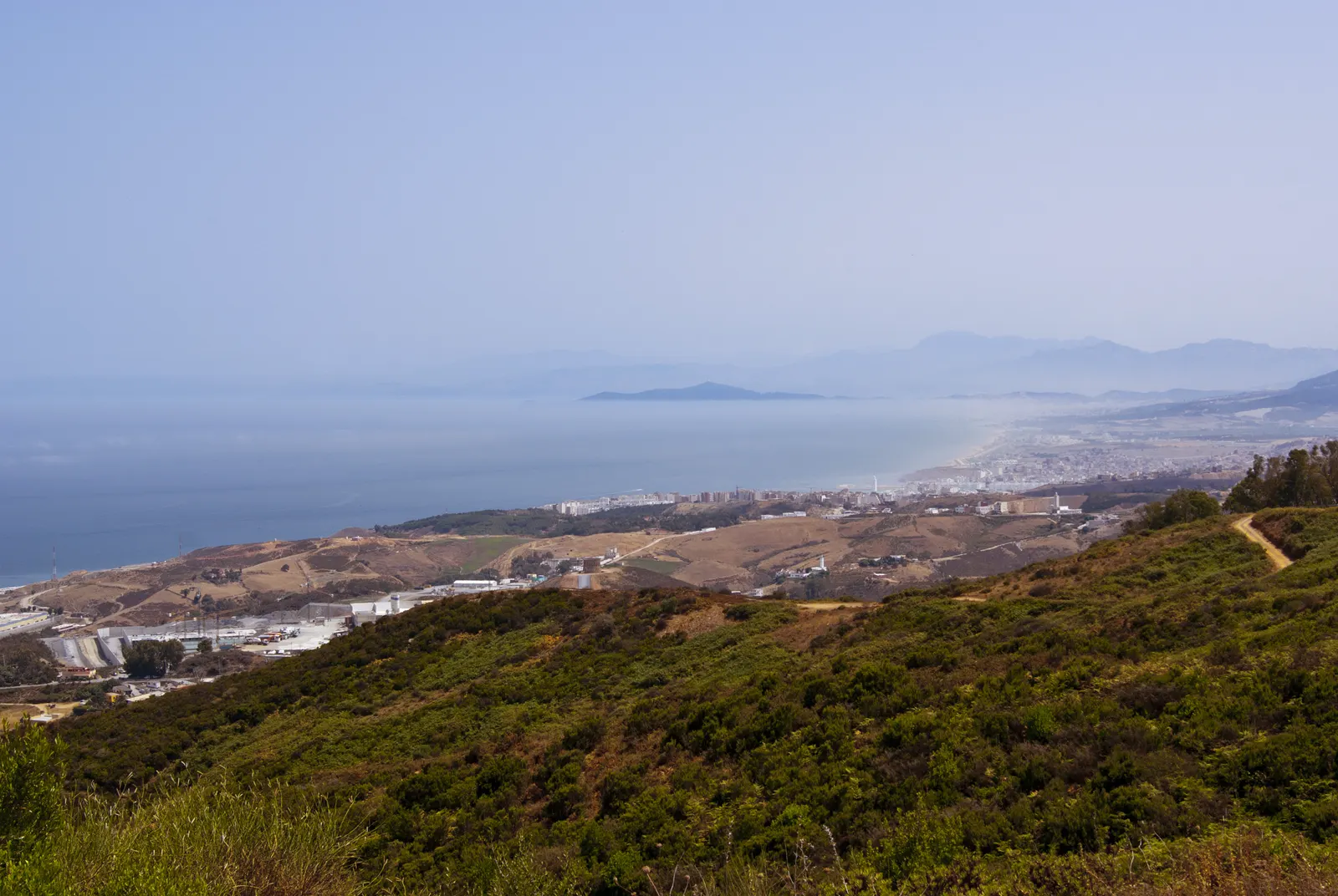 Vista desde los montes de Ceuta hacia el sur: laderas cubiertas de matorral en primer término, el mar a la izquierda y, al fondo, el caserío costero de Fnideq, en Marruecos, difuminado por la calima