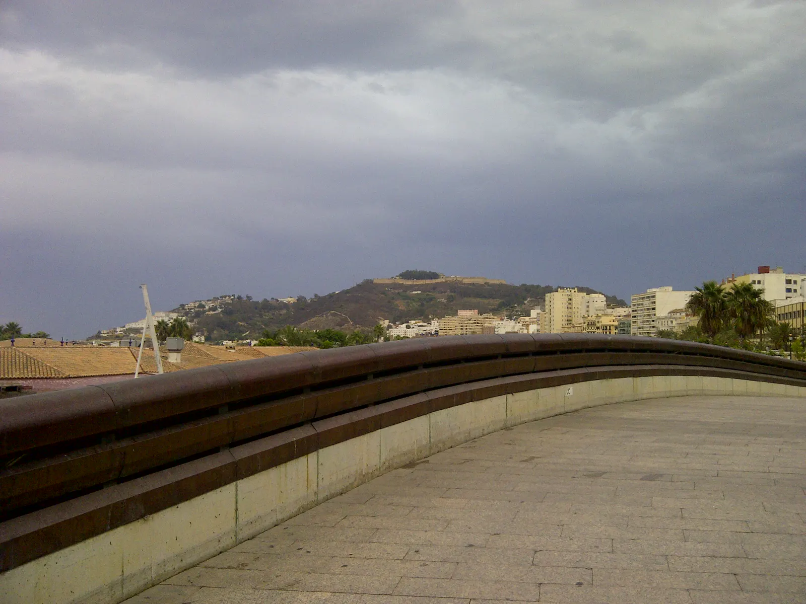 Vista de Ceuta desde el paseo marítimo: explanada con barandilla curva en primer término, tejados y edificios de la ciudad detrás y el monte Hacho con su fortaleza al fondo, bajo un cielo cubierto