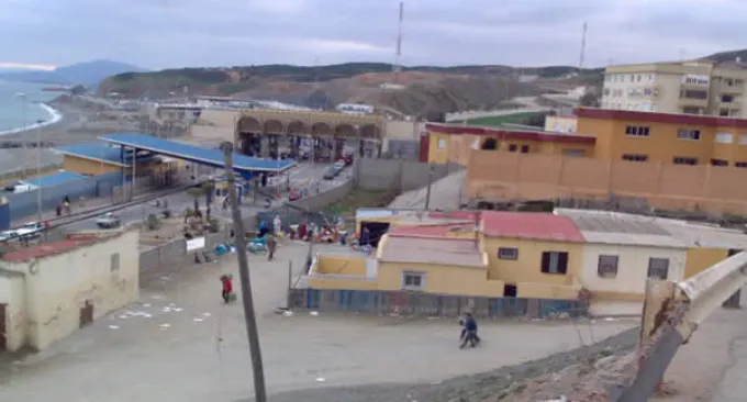 Vista elevada de la zona fronteriza de El Tarajal, en Ceuta: instalaciones del paso fronterizo junto al mar, con la playa y la costa curvándose hacia Marruecos al fondo bajo un cielo nublado