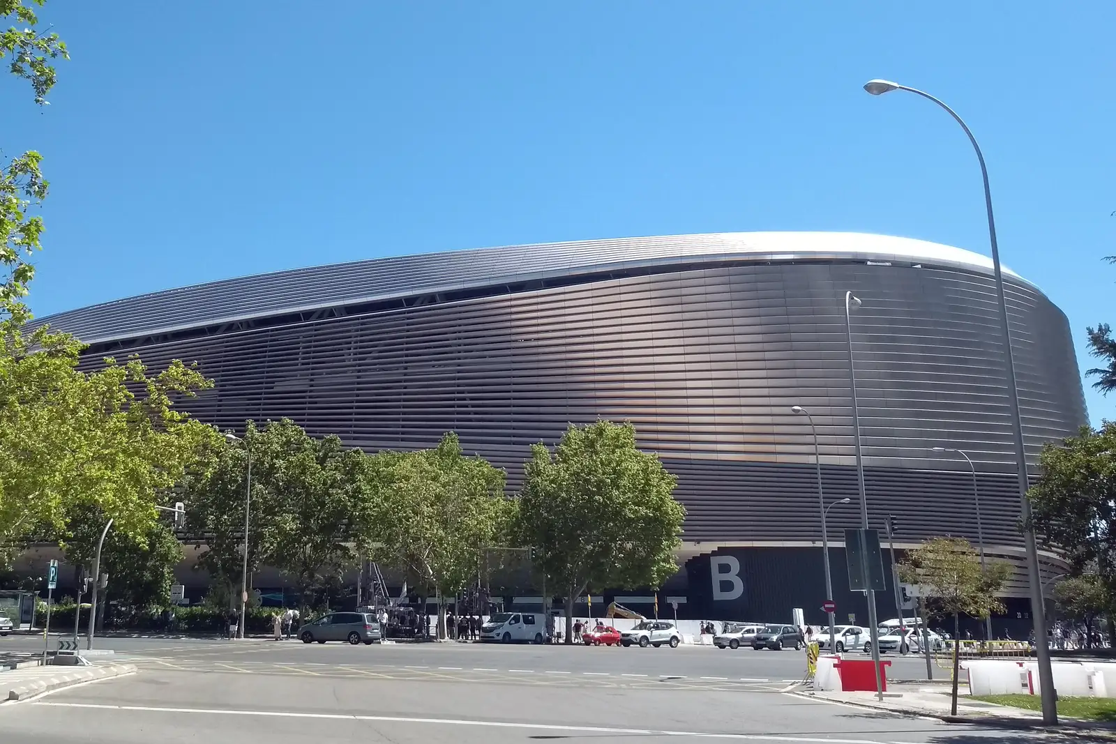 Fachada exterior del estadio Santiago Bernabéu, en Madrid: la envolvente de lamas metálicas curvas bajo un cielo despejado, con árboles, tráfico y viandantes a pie de calle