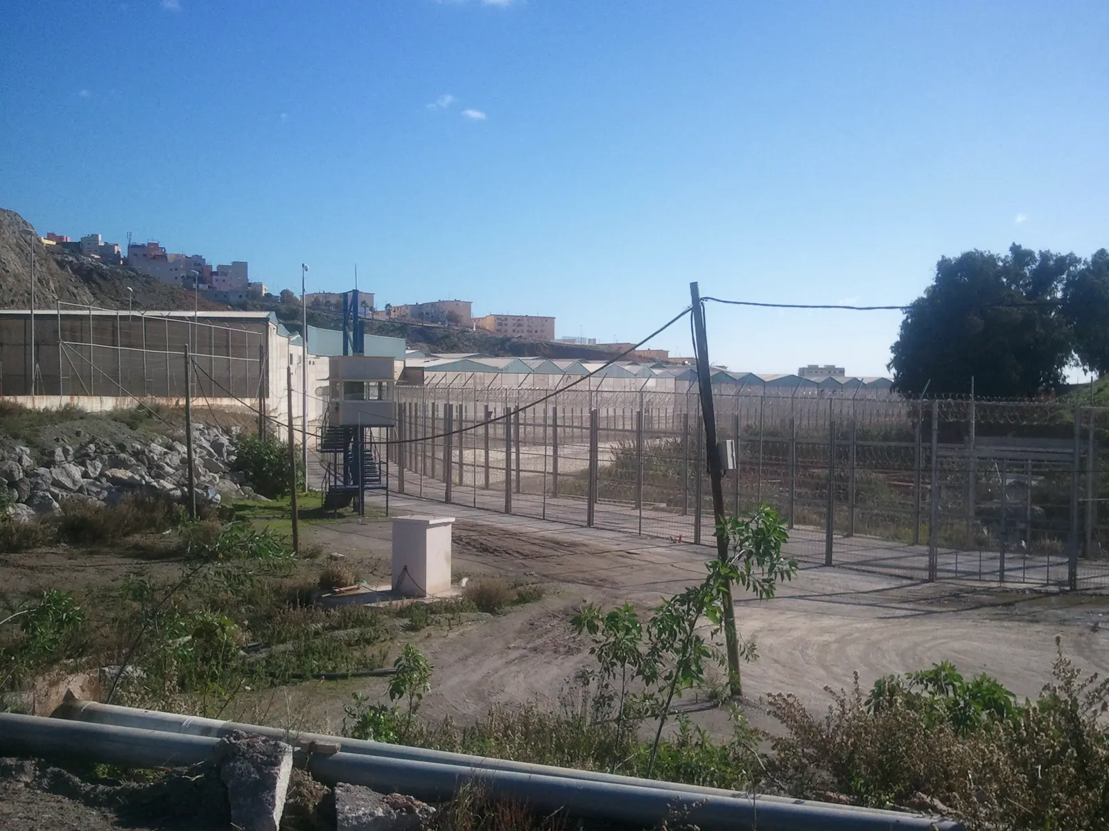 Valla fronteriza de Ceuta: doble hilera de vallas metálicas coronadas con concertinas, una torreta de vigilancia con escaleras y, al fondo, viviendas en la ladera del lado marroquí bajo un cielo despejado