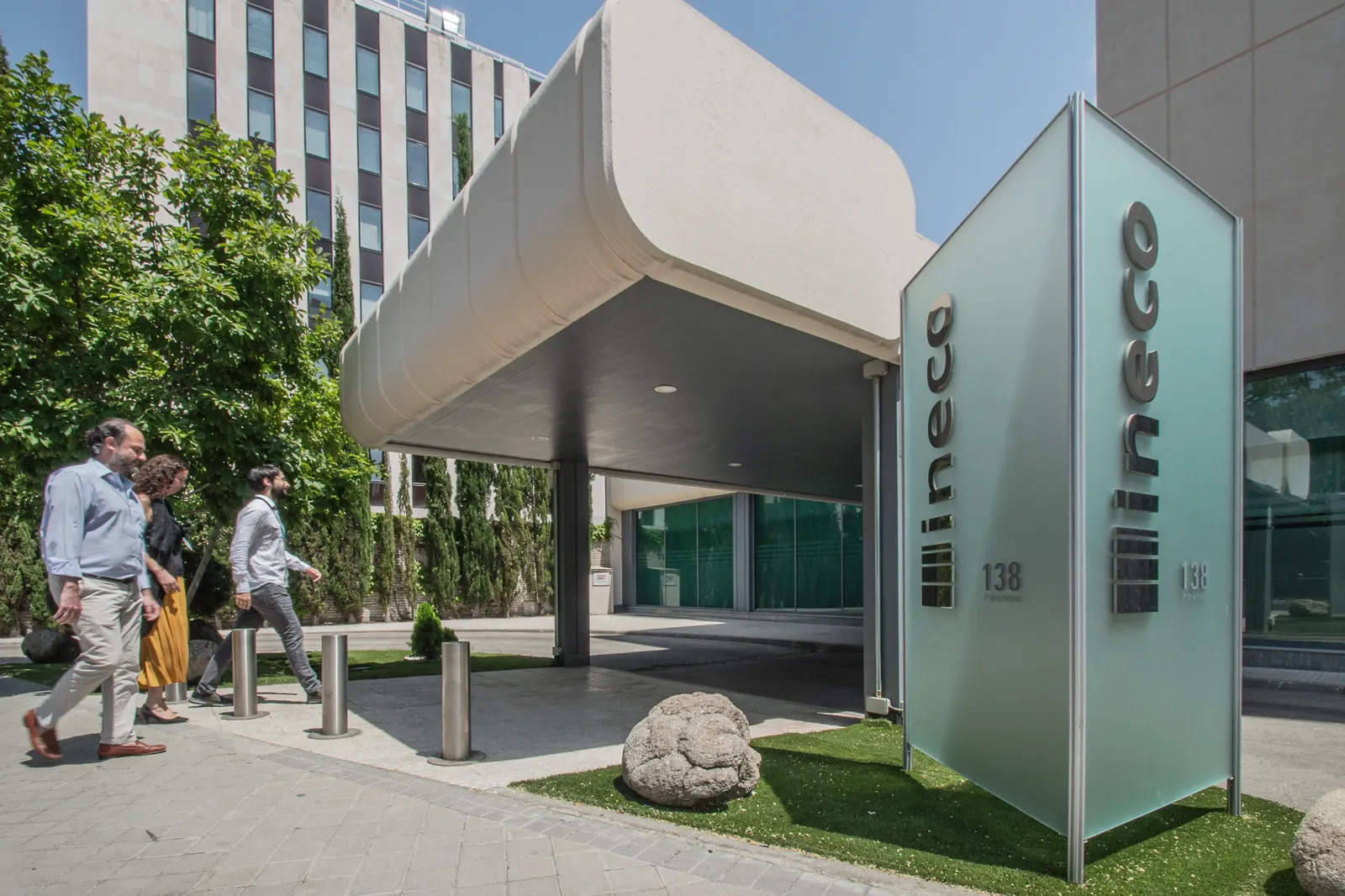Entrada de la sede central de Ineco en Madrid, con una marquesina blanca, un tótem de cristal con el logotipo de la empresa y el número 138, y tres personas caminando por la acera.