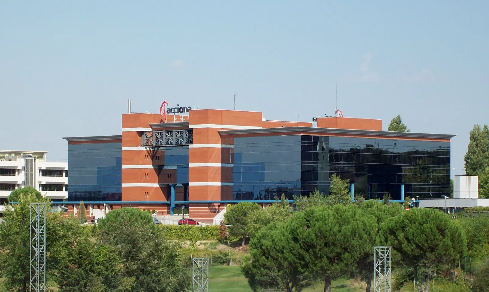 Edificio corporativo de ladrillo rojo y cristal oscuro rodeado de pinos, con el rótulo y el logotipo de Acciona en la cubierta, bajo un cielo despejado.