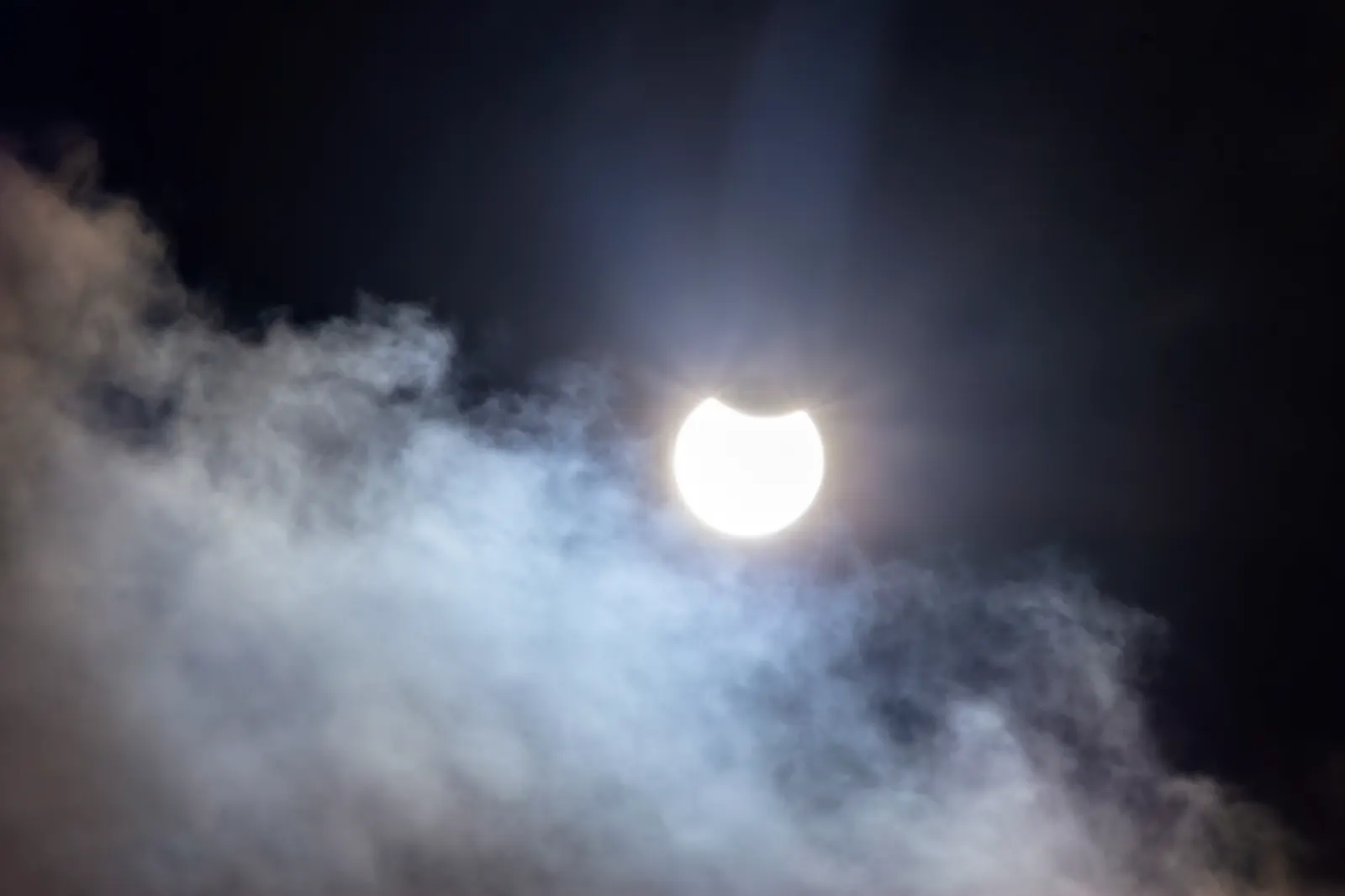 El Sol, con un mordisco oscuro en su borde superior por el paso de la Luna, brilla entre nubes recortadas sobre un cielo casi negro