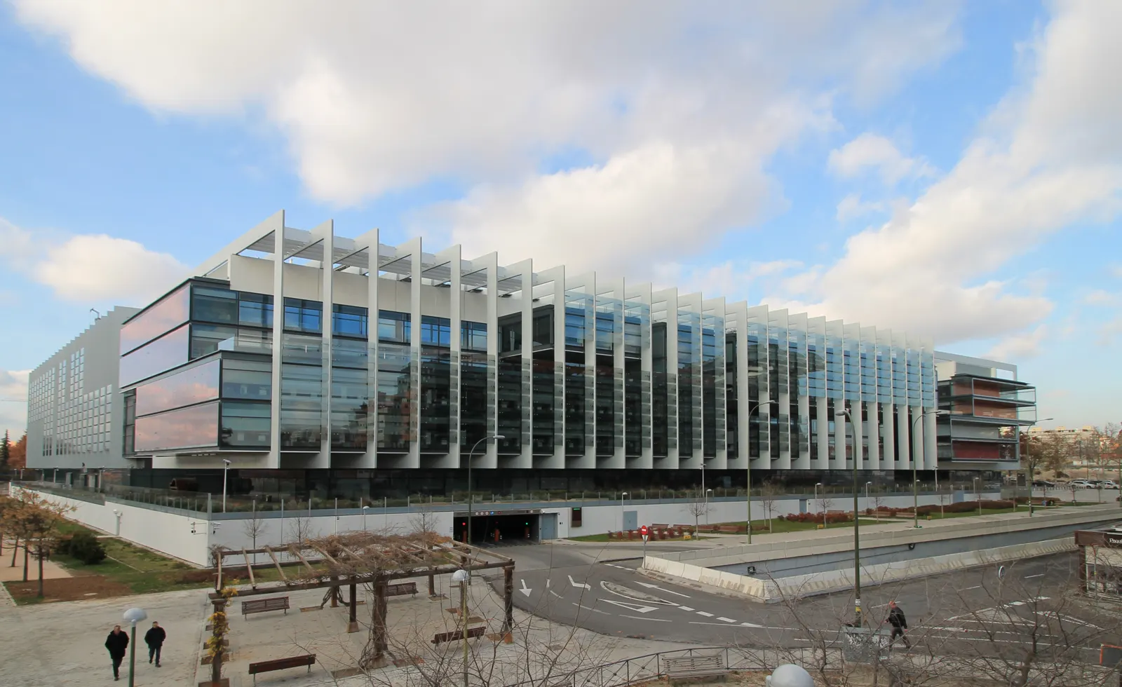 Fachada sureste del Campus Repsol en Madrid: edificio de vidrio y celosías blancas dispuesto en torno a un patio, con árboles jóvenes en primer término y cielo despejado.