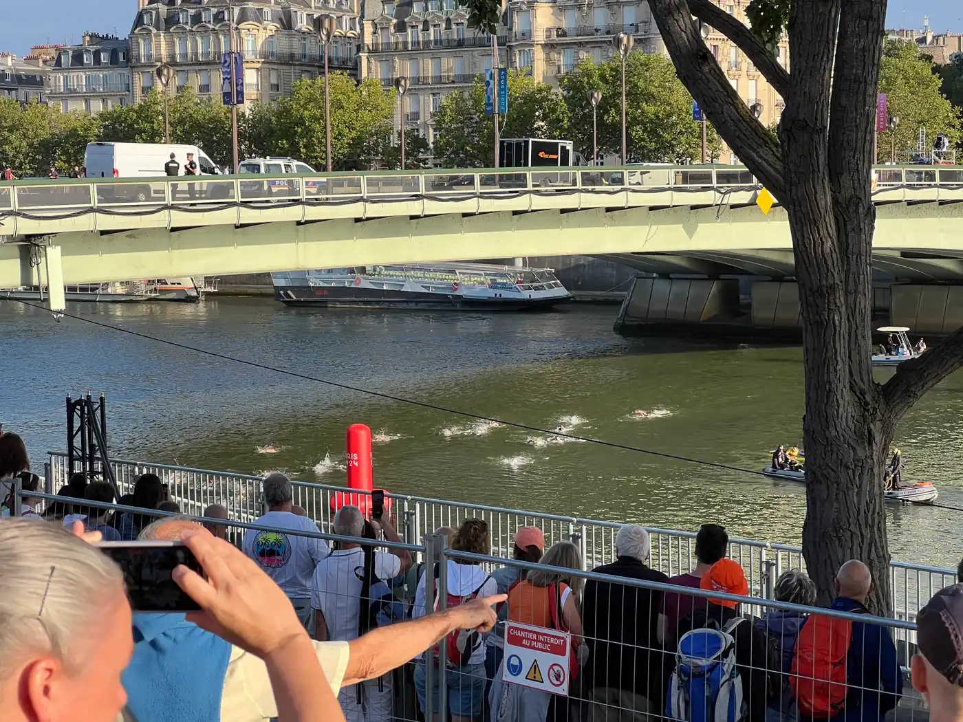 Varias nadadoras avanzan por el Sena junto a una boya roja de viraje, mientras el público las sigue desde el muelle tras una valla y un puente metálico cruza el río al fondo.