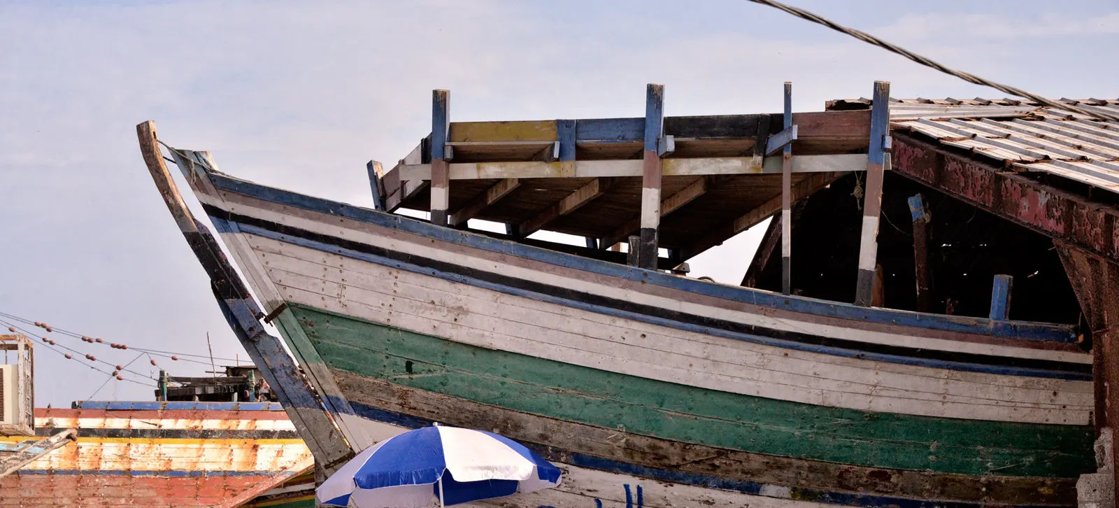 Casco de madera de un gran barco tradicional apuntalado en seco en un varadero, con las cuadernas y la cubierta a la vista y otras embarcaciones de madera al fondo.