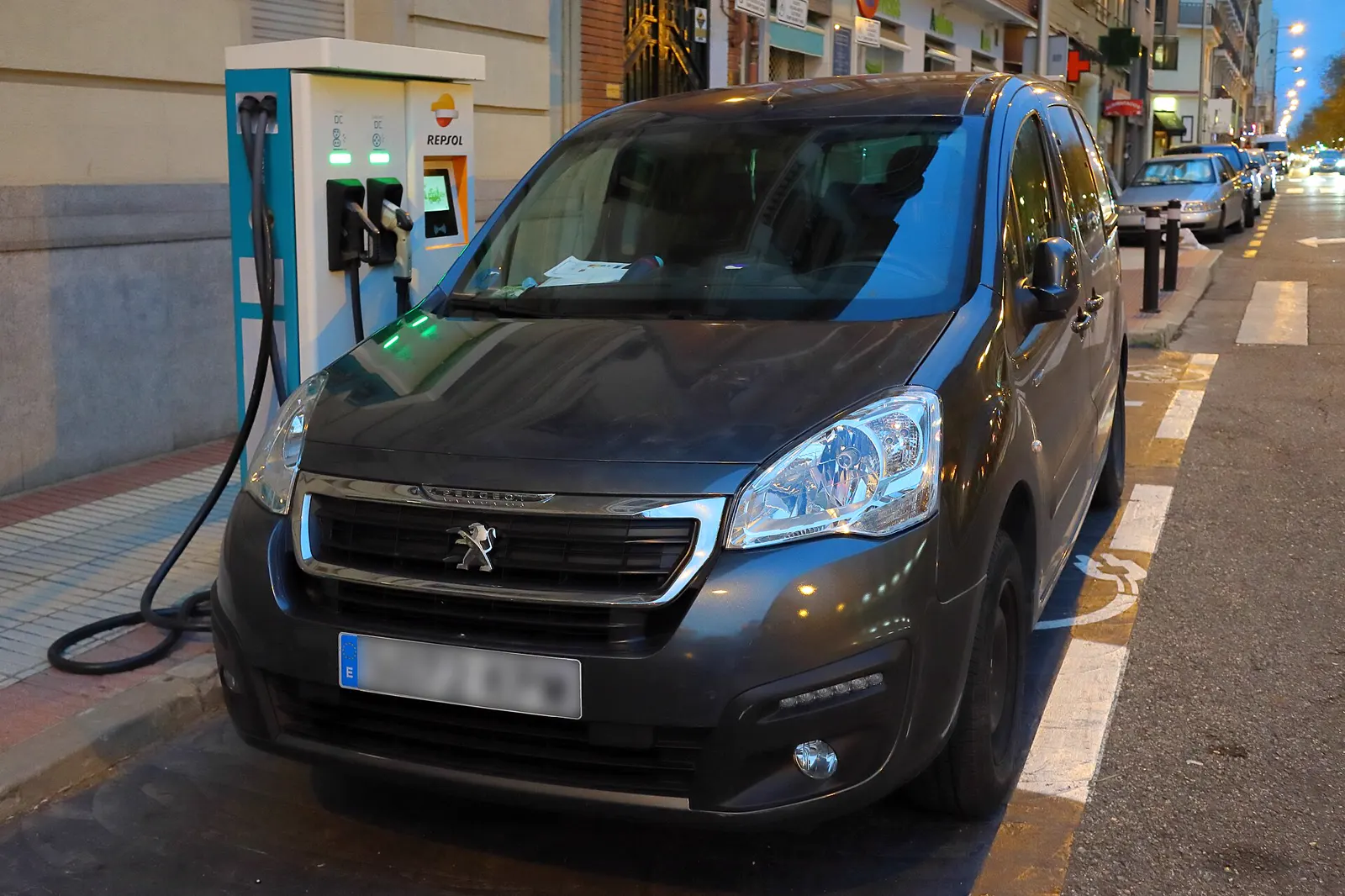 Furgoneta eléctrica Peugeot Partner enchufada a un poste de recarga en una calle de Madrid al anochecer.