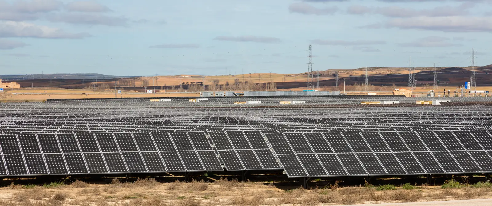 Filas de paneles solares fotovoltaicos cubren una llanura de campos de secano, con torres de alta tensión y cerros al fondo bajo un cielo nuboso.