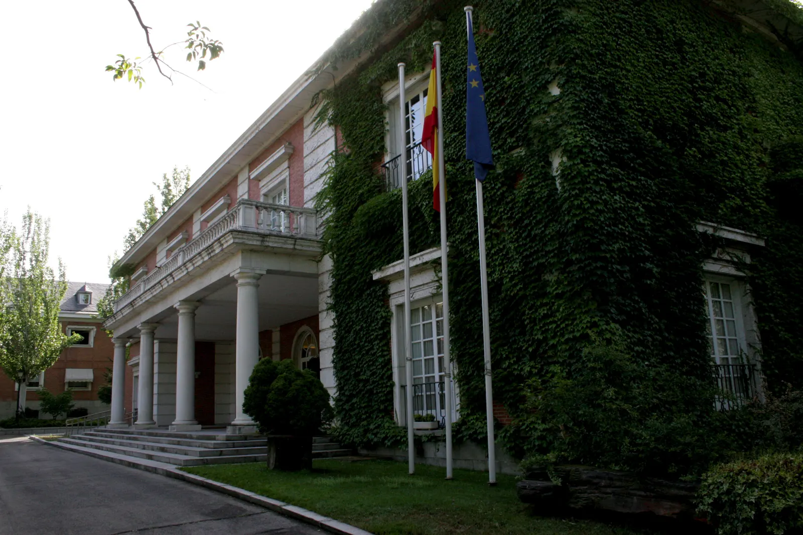 Edificio de ladrillo cubierto de hiedra con un pórtico de columnas blancas y una escalinata de acceso; ante la fachada ondean la bandera de España y la de la Unión Europea
