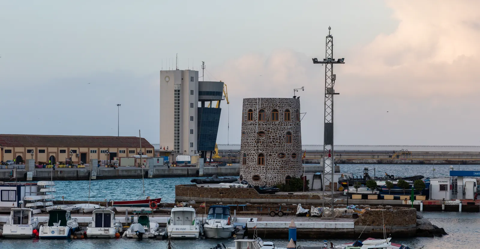Vista del puerto de Ceuta al atardecer: barcas amarradas en primer término, un muelle con naves industriales y una grúa, una torre de piedra junto al agua y el faro de una torre de control al fondo