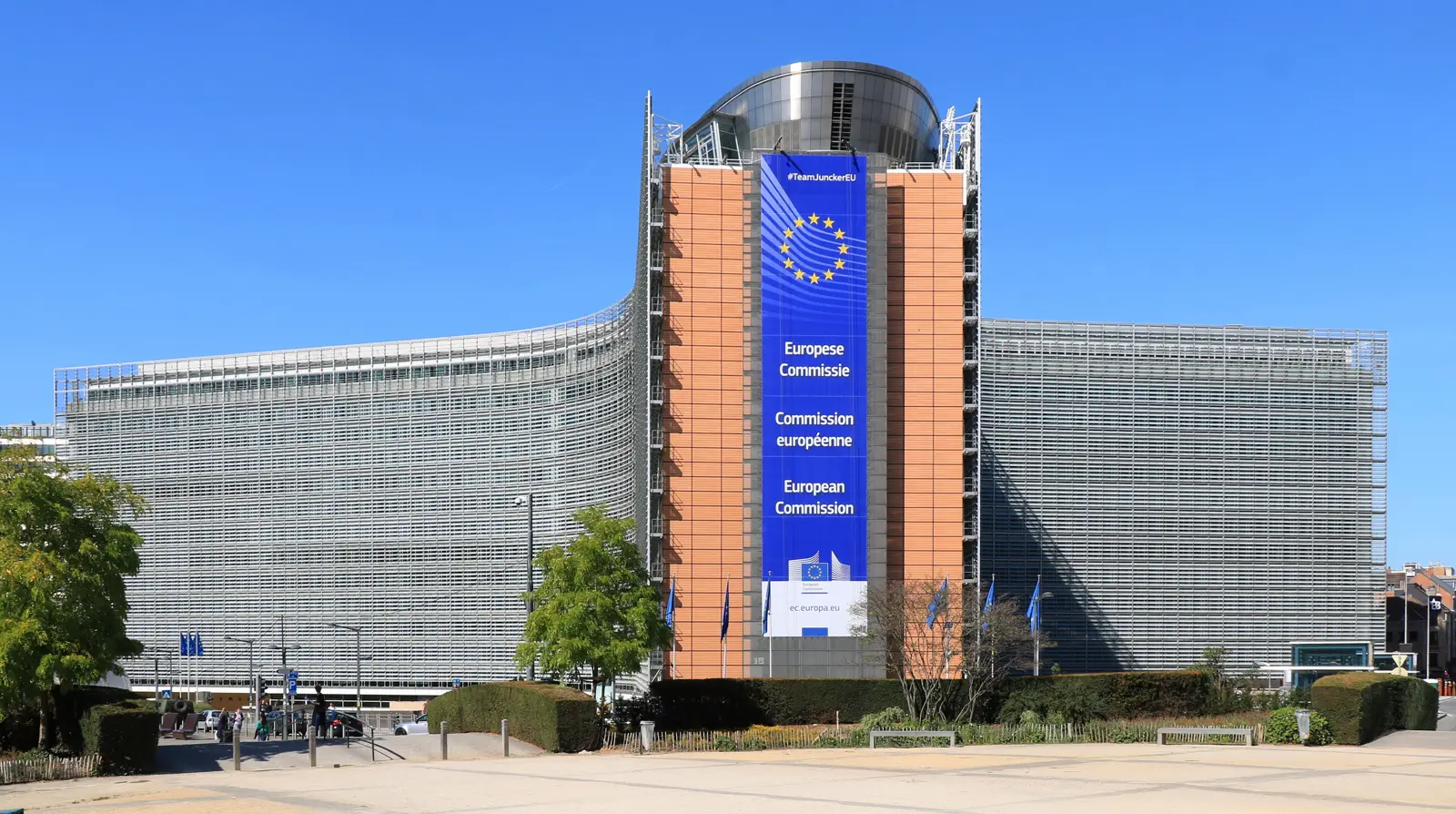 Fachada curva del edificio Berlaymont, sede de la Comisión Europea en Bruselas, con un gran cartel azul con las estrellas de la Unión Europea y el rótulo «European Commission» colgado de la torre central, banderas europeas a pie de calle y cielo despejado.