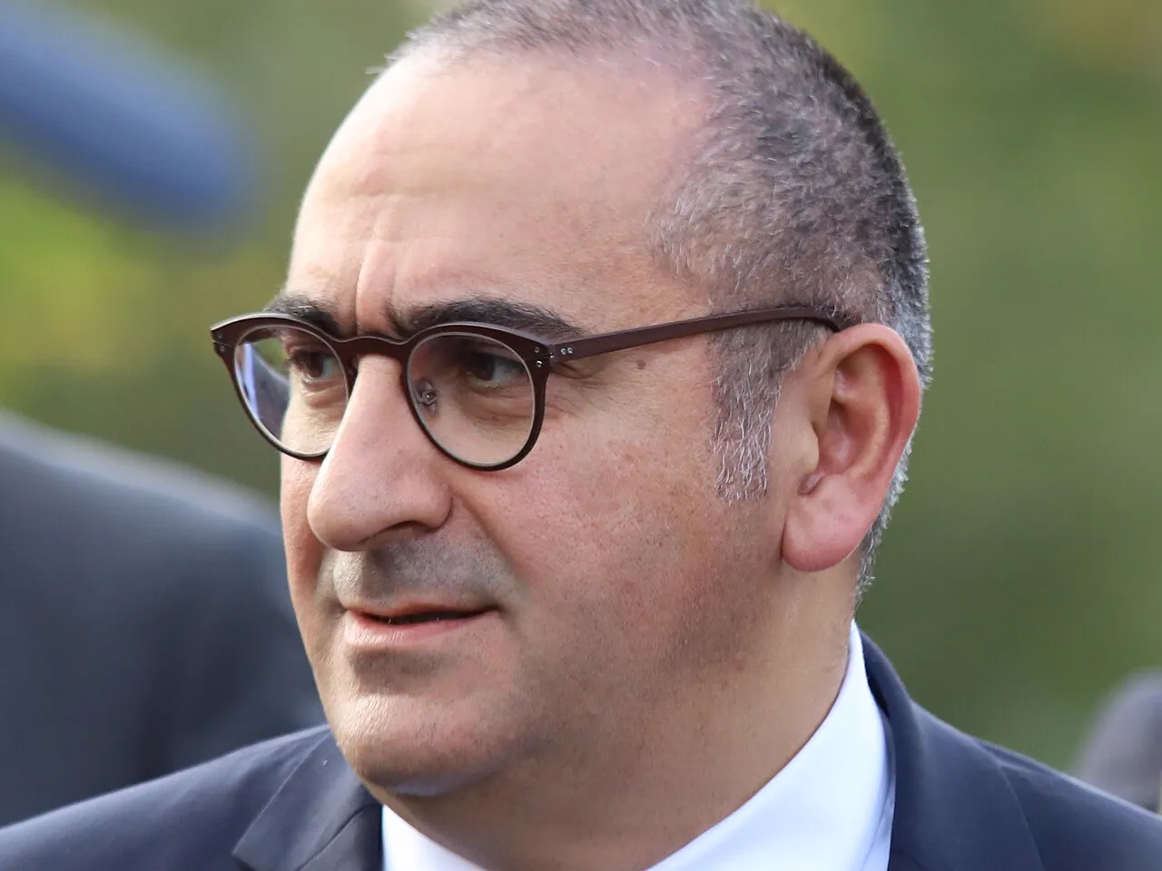 Retrato de un hombre de mediana edad con gafas redondas de montura oscura, pelo corto y canoso, traje azul marino, camisa blanca y corbata azul, al aire libre y con vegetación desenfocada al fondo.