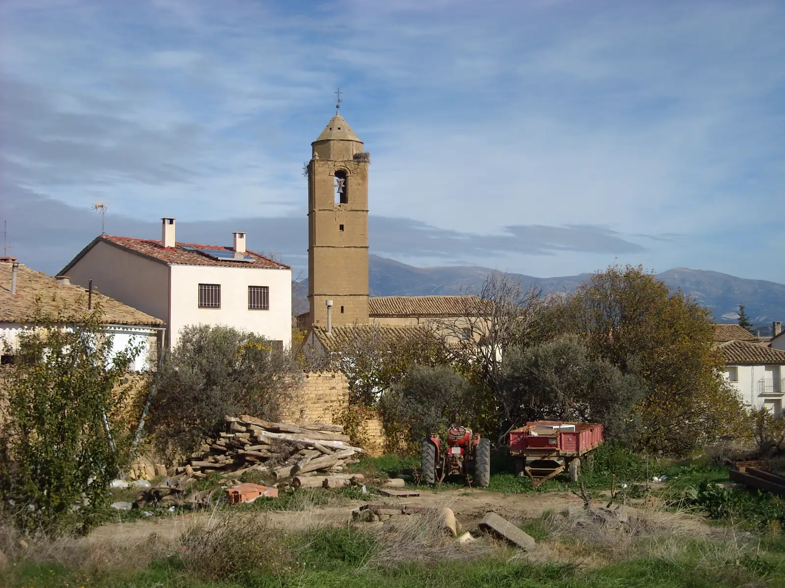 Vista del pueblo de Loporzano (Huesca): la torre octogonal de piedra de la iglesia sobresale entre casas encaladas y tejados rojos; en primer plano, un huerto con un tractor rojo y un remolque junto a un montón de leña, y al fondo, la línea azulada de la sierra.