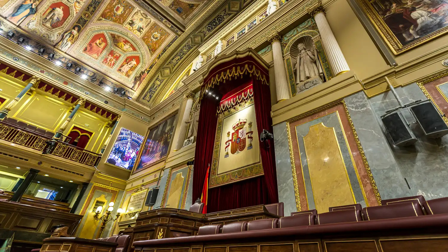 Hemiciclo vacío del Congreso de los Diputados visto desde los escaños: el tapiz con el escudo de España sobre la tribuna, techos artesonados con pinturas y bancada de escaños de madera y cuero