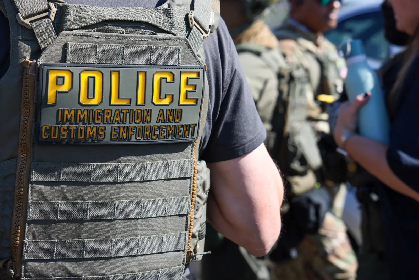 Primer plano del chaleco táctico de un agente, con un parche amarillo y negro que dice «Police - Immigration and Customs Enforcement». Al fondo, desenfocados, otros agentes de uniforme.