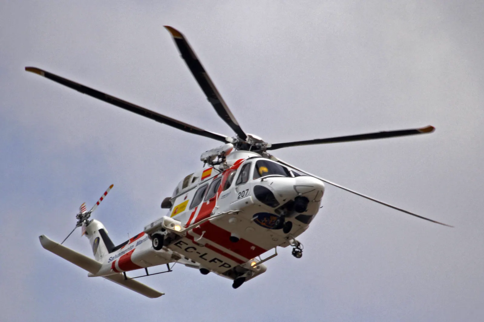 Helicóptero de Salvamento Marítimo, blanco con franjas rojas y matrícula EC-LFP, volando con el tren de aterrizaje desplegado bajo un cielo gris