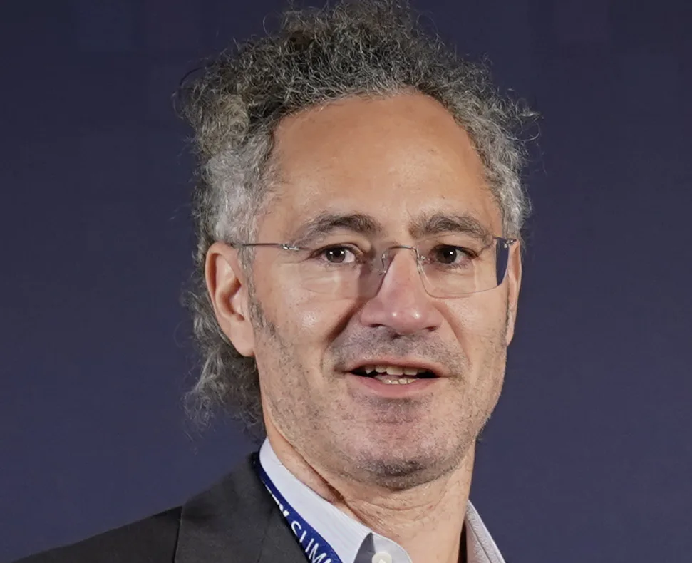Retrato de Alex Karp, hombre de pelo cano y rizado con gafas metálicas y traje oscuro, con una acreditación azul de la cumbre de seguridad de la inteligencia artificial colgada al cuello