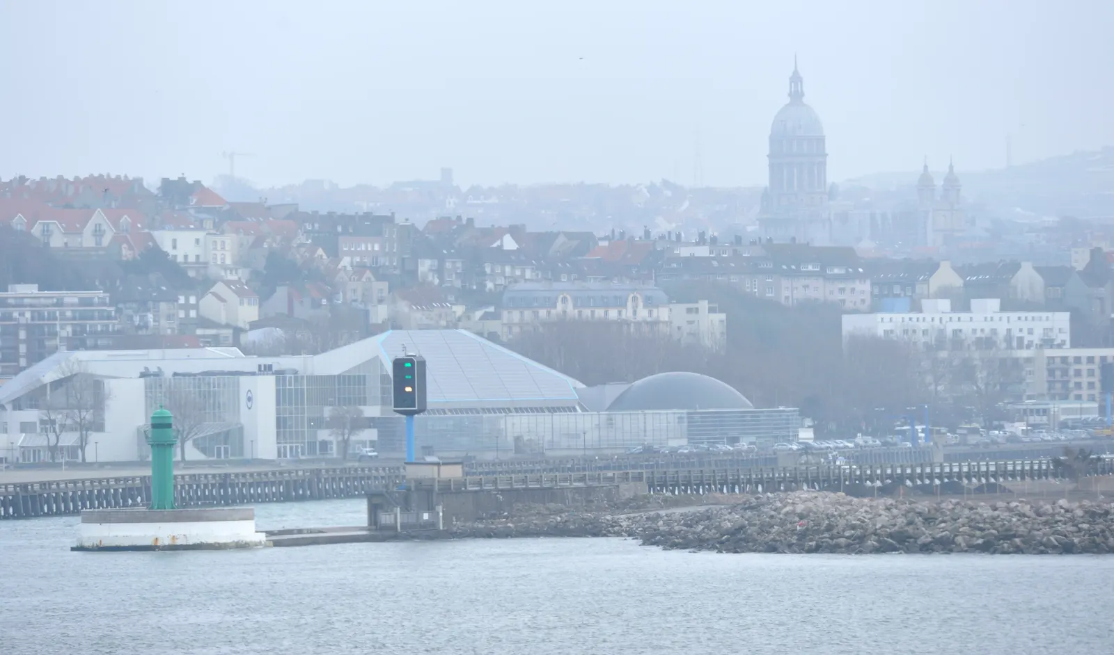 Entrada del puerto de Boulogne-sur-Mer vista desde el mar, con una baliza verde sobre el espigón y, al fondo entre la bruma, la ciudad y la cúpula de la basílica de Notre-Dame