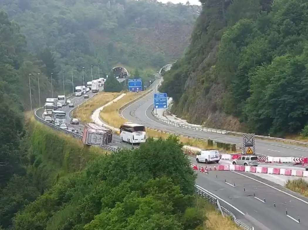 Vista elevada de una autovía entre laderas boscosas: un camión volcado sobre la calzada, una larga cola de camiones y coches detenidos detrás y, en el otro sentido, conos y balizas que estrechan los carriles.