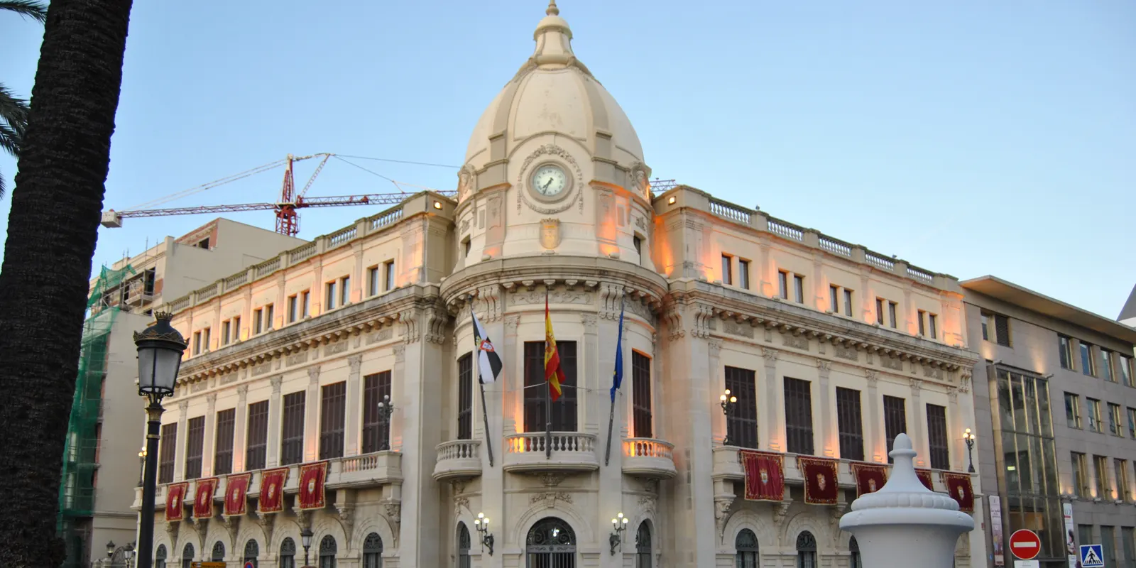 Fachada del Palacio de la Asamblea de Ceuta al atardecer: edificio de piedra clara con cúpula y reloj sobre el chaflán, balcón con las banderas de Ceuta, España y la Unión Europea, y una grúa de obra al fondo.