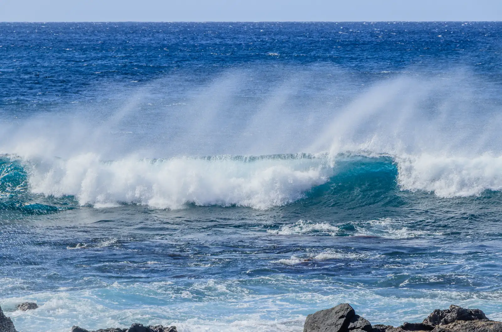 Una ola rompe frente a una costa de rocas negras y el viento arrastra hacia atrás la espuma en penachos blancos sobre un mar azul intenso.