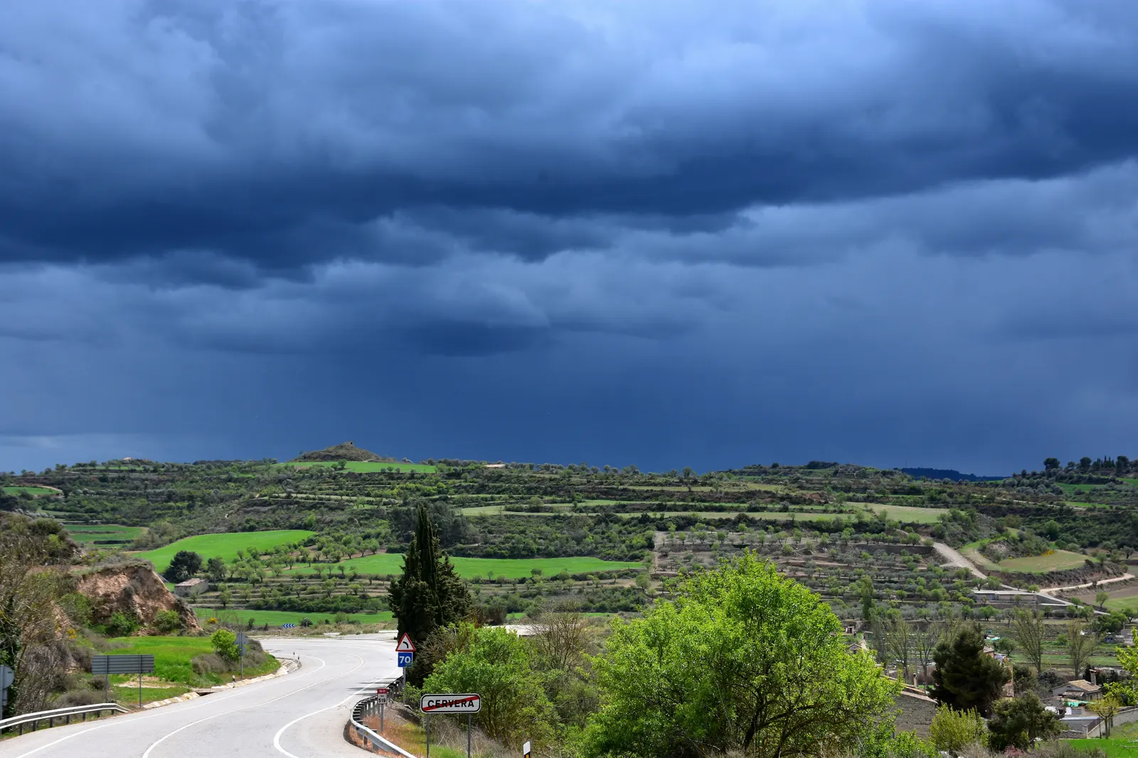 Un frente de nubes de tormenta muy oscuras cubre el cielo sobre campos verdes, olivos y bancales de la Segarra; en primer plano, una carretera con un cartel que dice «Cervera».