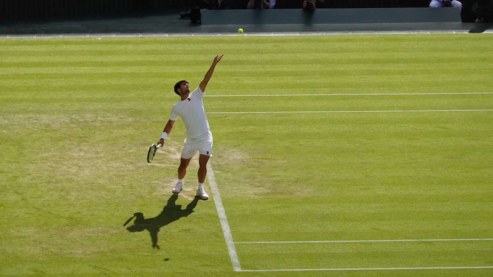 Carlos Alcaraz, vestido de blanco, en pleno gesto de saque sobre la hierba de una pista de Wimbledon: el brazo izquierdo alzado tras lanzar la pelota, que flota en el aire, y la raqueta en la mano derecha, a la espalda.