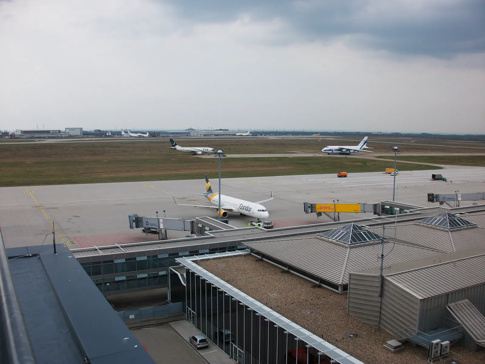 Vista elevada del campo de vuelo del aeropuerto de Leipzig/Halle, con un avión de pasajeros de Condor en plataforma, un carguero Antonov An-124 rodando al fondo y una furgoneta amarilla de DHL junto a la terminal.