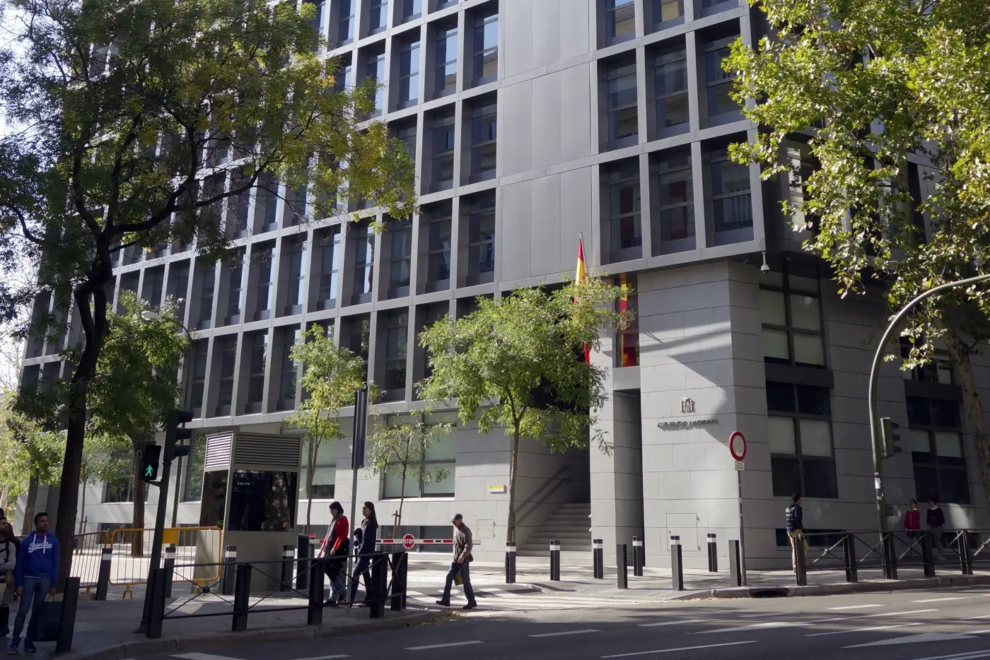 Fachada de una sede de la Audiencia Nacional en Madrid: edificio de hormigón y vidrio con ventanas verticales estrechas, una bandera de España sobre la entrada y varios peatones cruzando la acera arbolada.