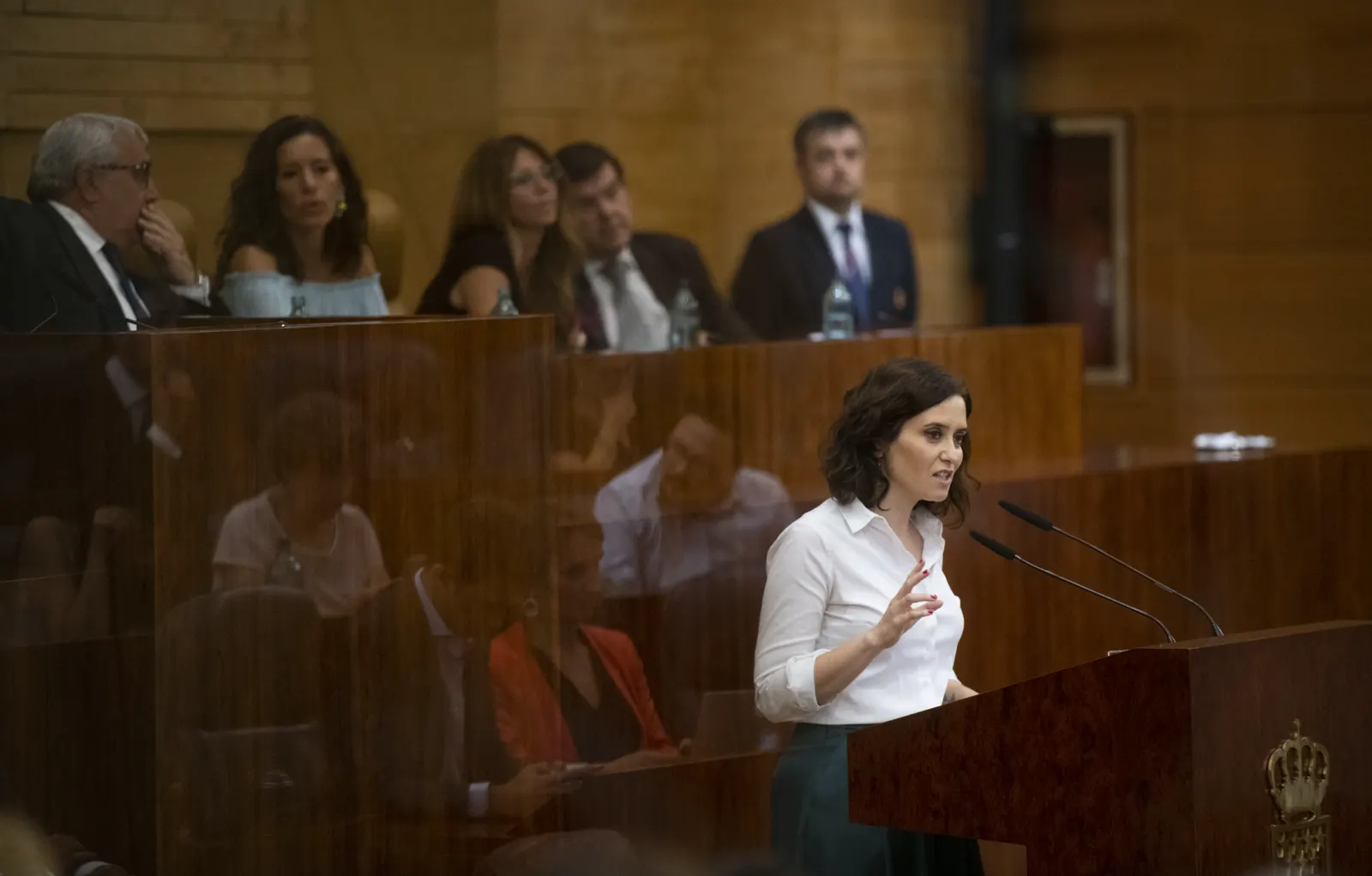 Isabel Díaz Ayuso habla ante dos micrófonos en la tribuna de madera de la Asamblea de Madrid, con la mesa de la presidencia y varios diputados al fondo