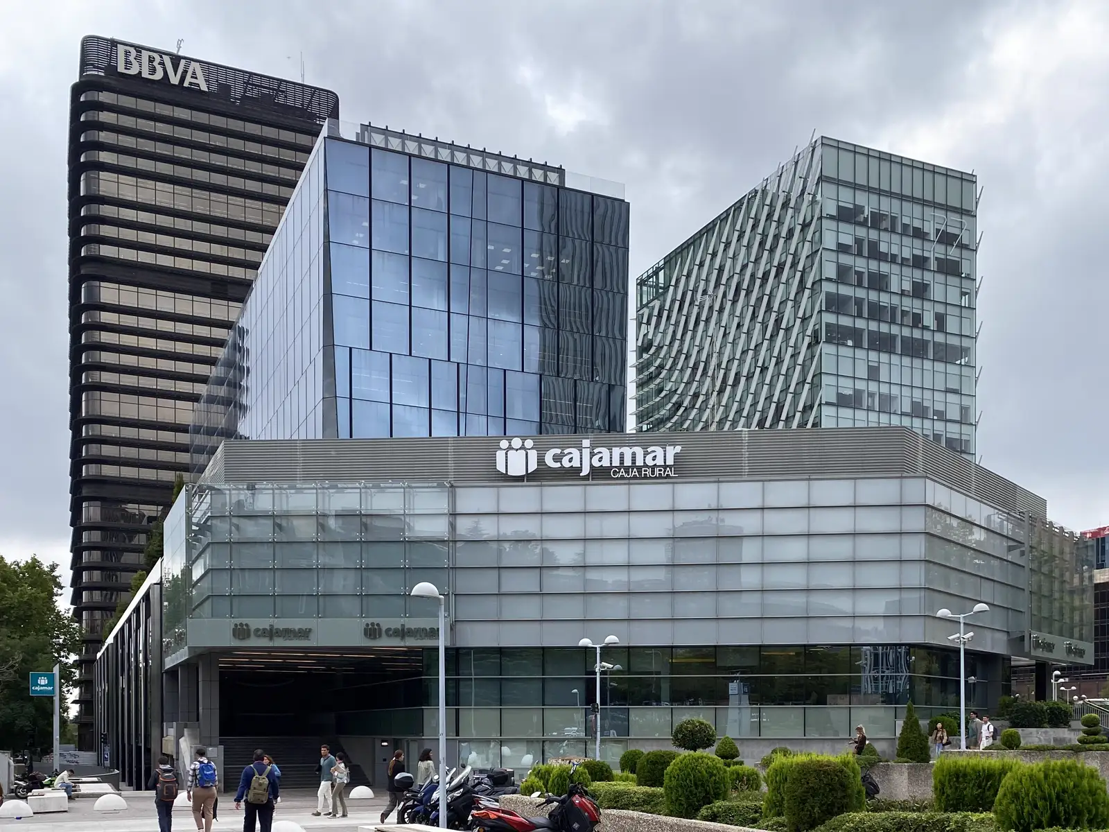 Edificio acristalado de oficinas con un gran rótulo de Cajamar Caja Rural sobre la entrada; detrás asoma la torre del BBVA y varios peatones cruzan la plaza