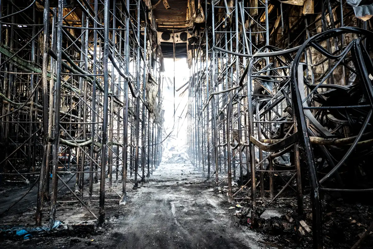 Interior de una nave logística calcinada, con las estanterías metálicas retorcidas por el fuego y el suelo cubierto de escombros y ceniza.
