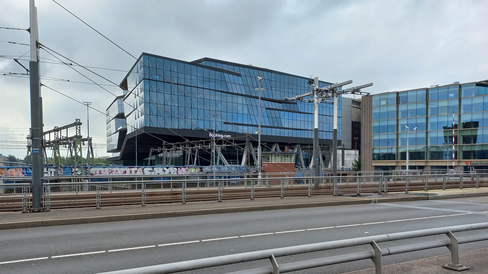 Edificio acristalado de planta inclinada con el rótulo «Booking.com» sobre la entrada, visto desde la otra orilla de una vía de tren, bajo un cielo nublado.