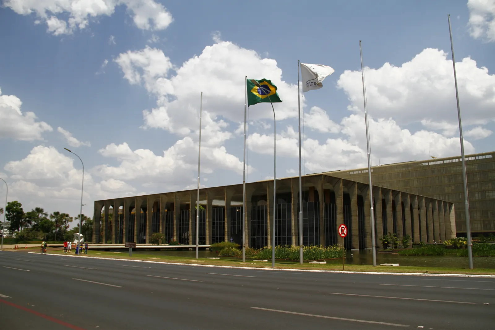 Edificio bajo y alargado de hormigón con una galería de arcos, visto desde una avenida vacía; delante, una hilera de mástiles en los que ondean la bandera de Brasil y una bandera blanca, bajo un cielo con nubes