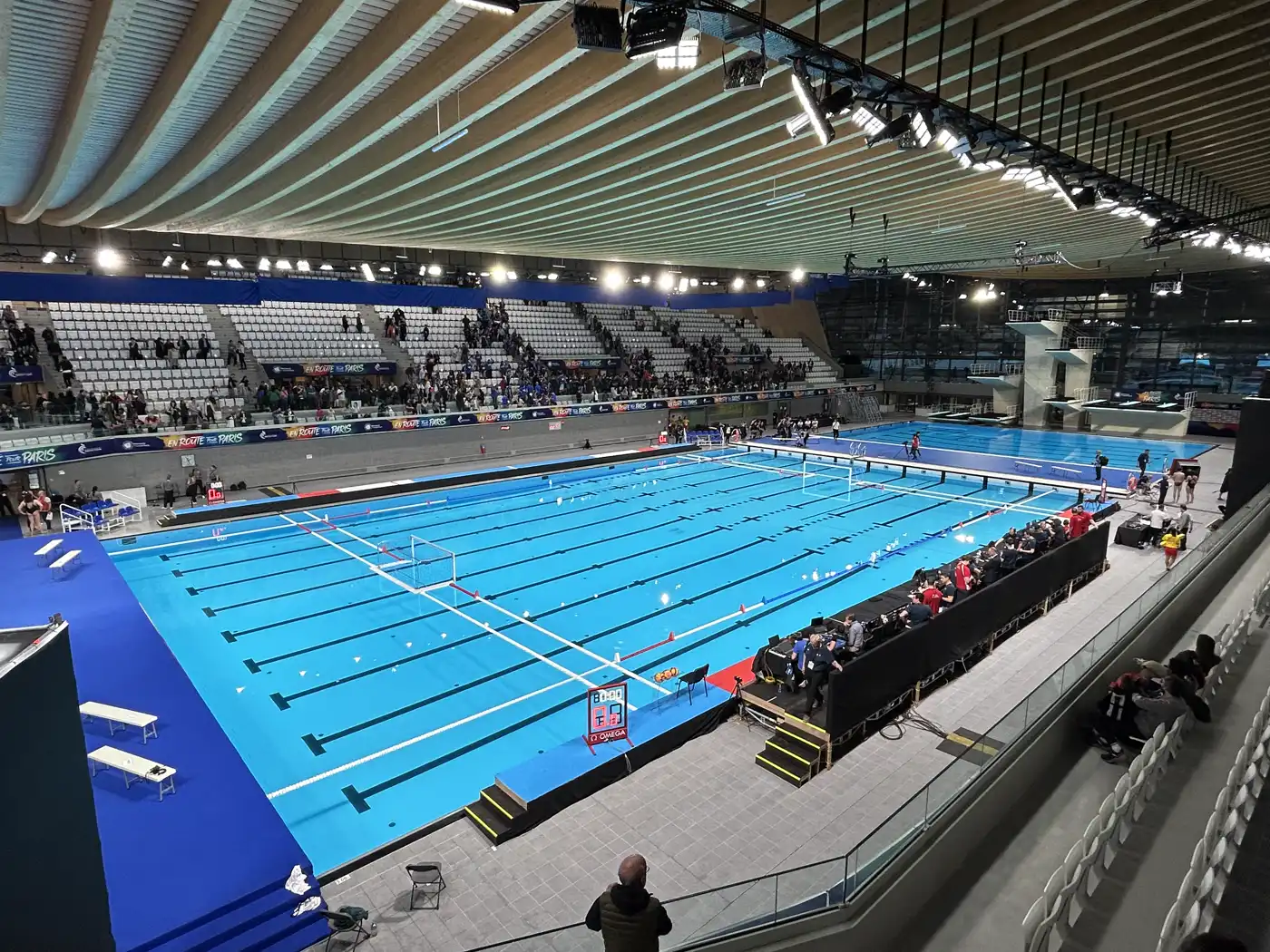 Vista desde la tribuna de la piscina del Centro Acuático Olímpico de Saint-Denis, con el techo curvo de madera, el graderío blanco parcialmente ocupado y, al fondo a la derecha, la torre blanca de plataformas de saltos junto al foso de saltos.