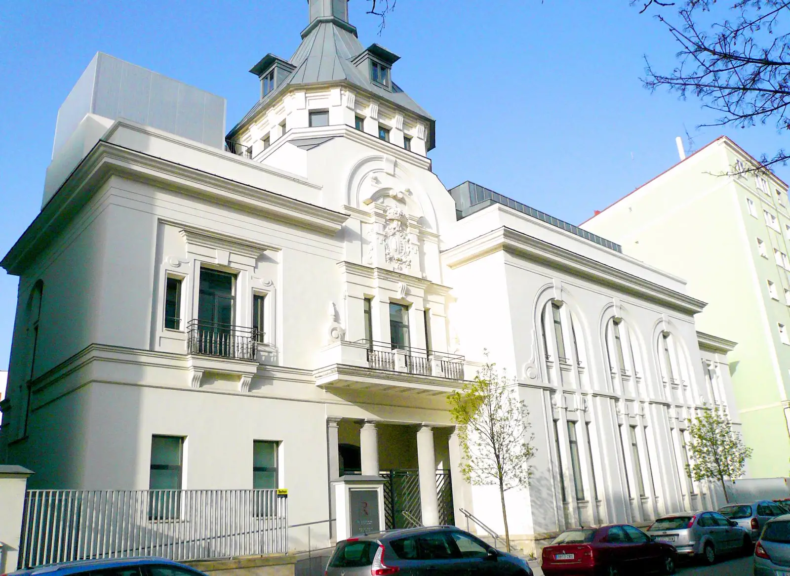 Edificio institucional de fachada blanca con torreón, escudo de piedra sobre el balcón central y pórtico de columnas en la entrada, con coches aparcados delante y cielo azul despejado.