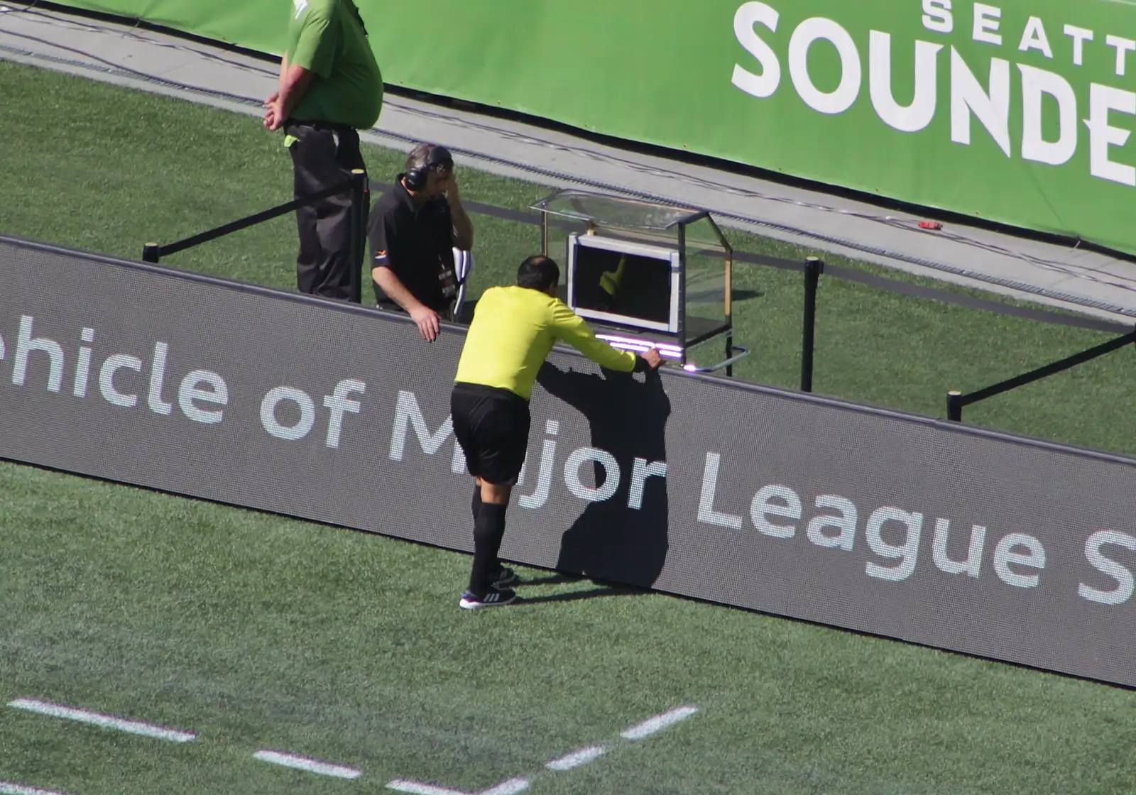 Un árbitro de fútbol, de espaldas y con camiseta amarilla, se inclina sobre el monitor del VAR instalado junto a la banda de un estadio de césped.