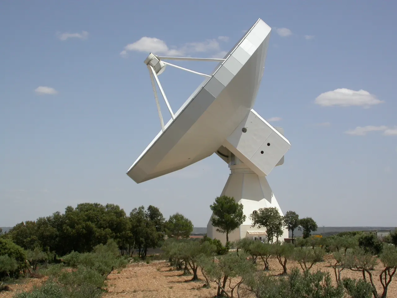Radiotelescopio blanco de gran tamaño, con la antena parabólica inclinada hacia el cielo, sobre un terreno seco poblado de olivos y encinas bajo un cielo azul con nubes dispersas
