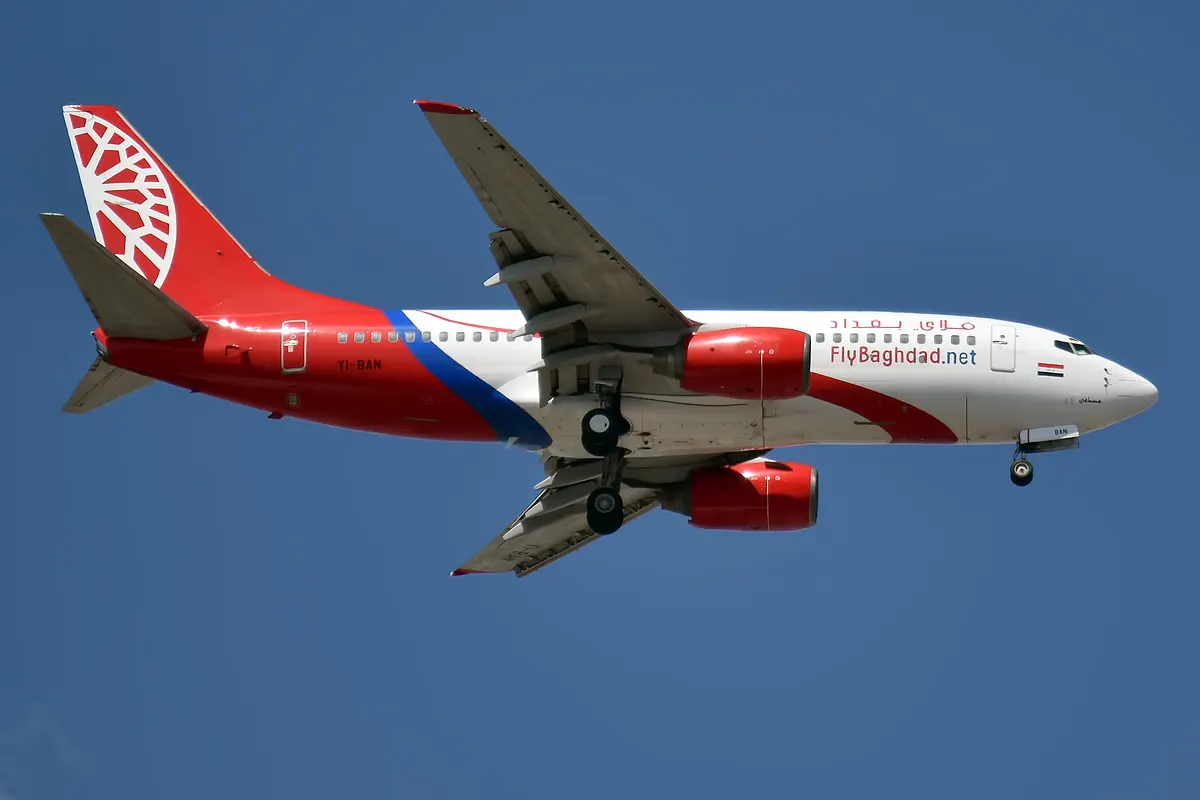 Avión Boeing 737 blanco y rojo de Fly Baghdad, con la matrícula YI-BAN pintada en el fuselaje, en vuelo con el tren de aterrizaje desplegado sobre un cielo azul despejado.