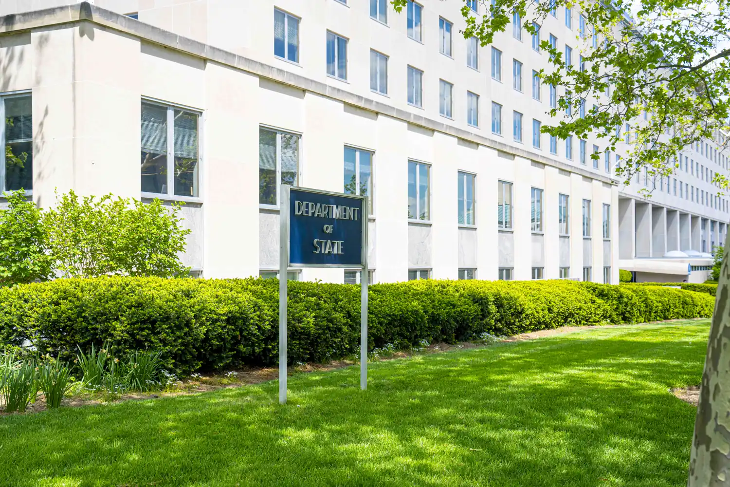 Fachada blanca del edificio Harry S. Truman, sede del Departamento de Estado en Washington, con un cartel metálico sobre césped en el que se lee «Department of State»
