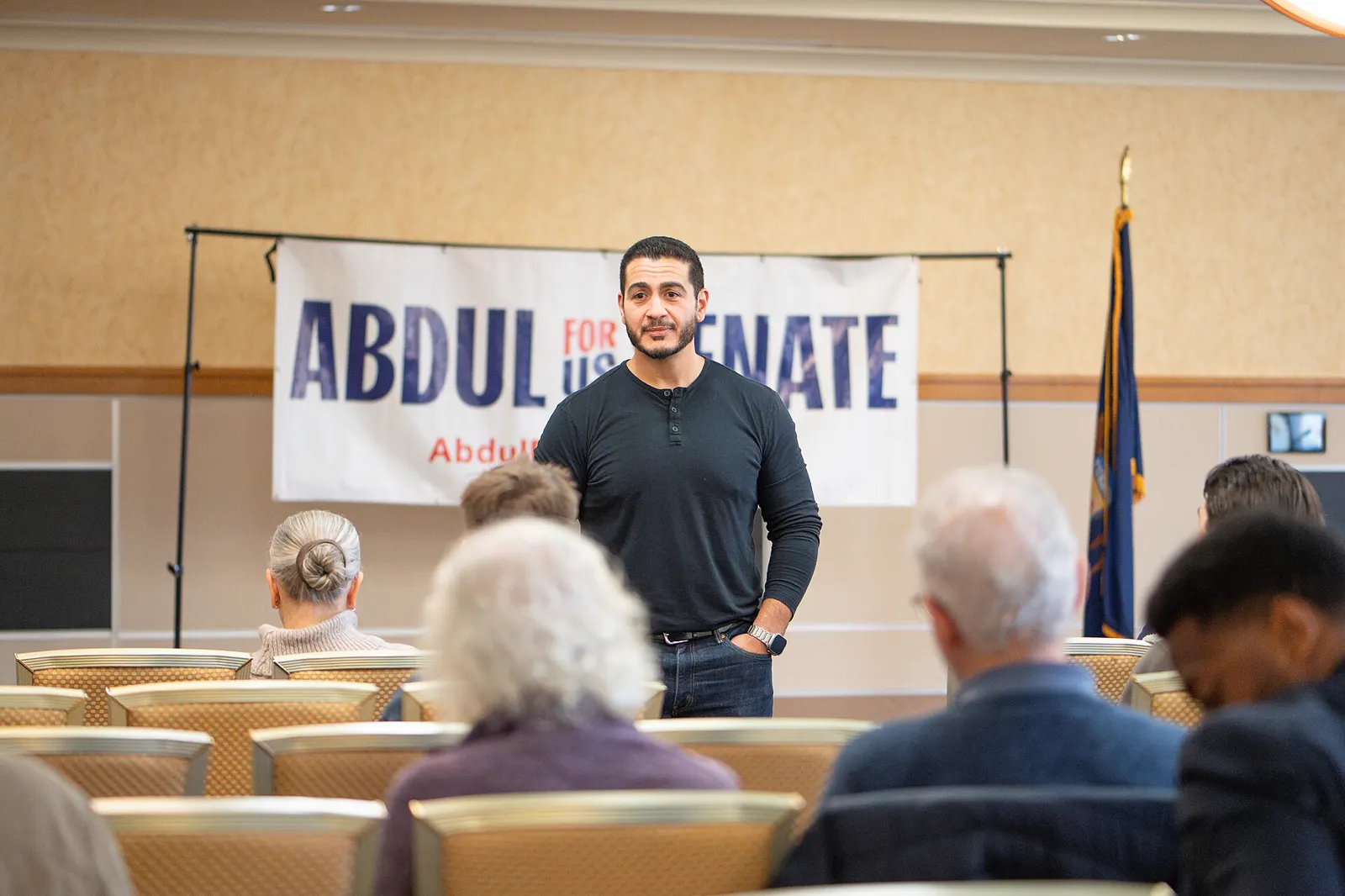 Abdul El-Sayed, con jersey oscuro, de pie ante una veintena de asistentes sentados, con una pancarta de campaña que dice «Abdul for US Senate» y una bandera de Michigan al fondo.