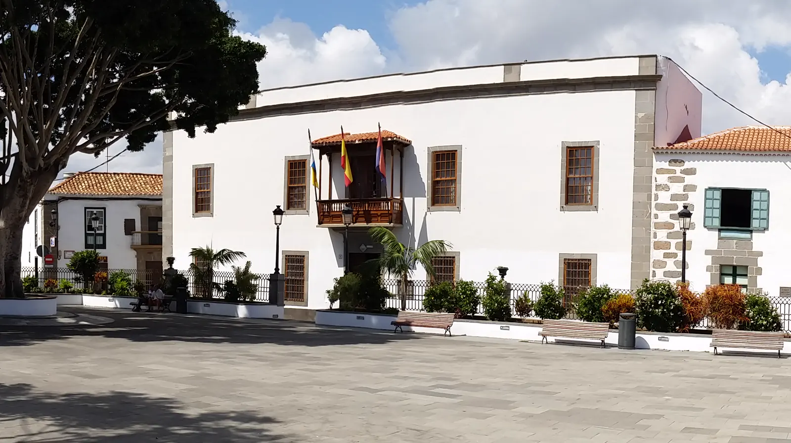 Fachada blanca de dos plantas con balcón de madera y tres banderas, frente a una plaza empedrada con bancos y un gran árbol.