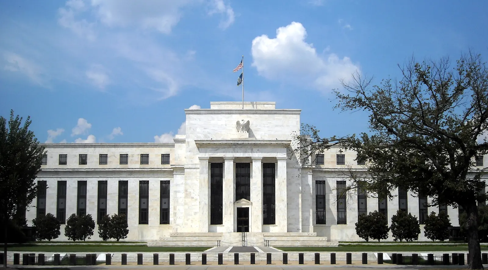 Fachada neoclásica de piedra blanca del edificio Marriner S. Eccles, sede del consejo de gobernadores de la Reserva Federal, en Washington, con árboles delante y cielo azul.