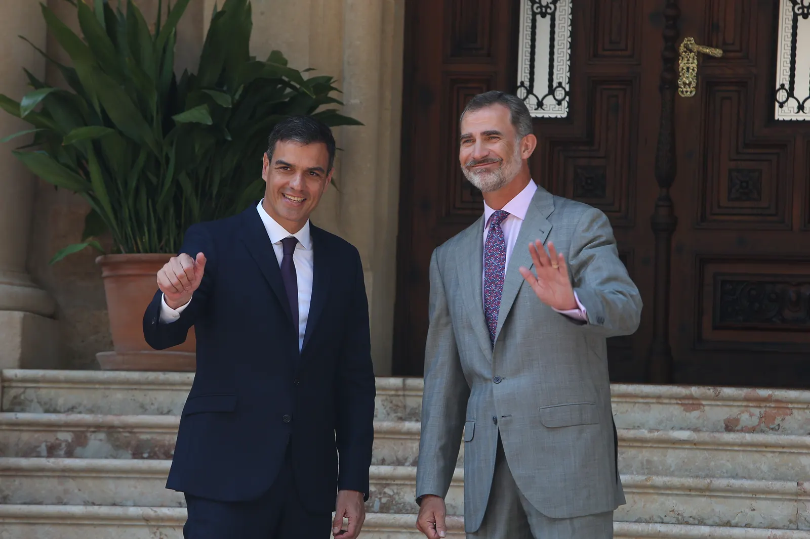 Felipe VI, con traje gris claro, saluda con la mano en lo alto de la escalinata del palacio de Marivent, junto al presidente del Gobierno, Pedro Sánchez, de traje oscuro, ante el portón de madera labrada de la entrada.