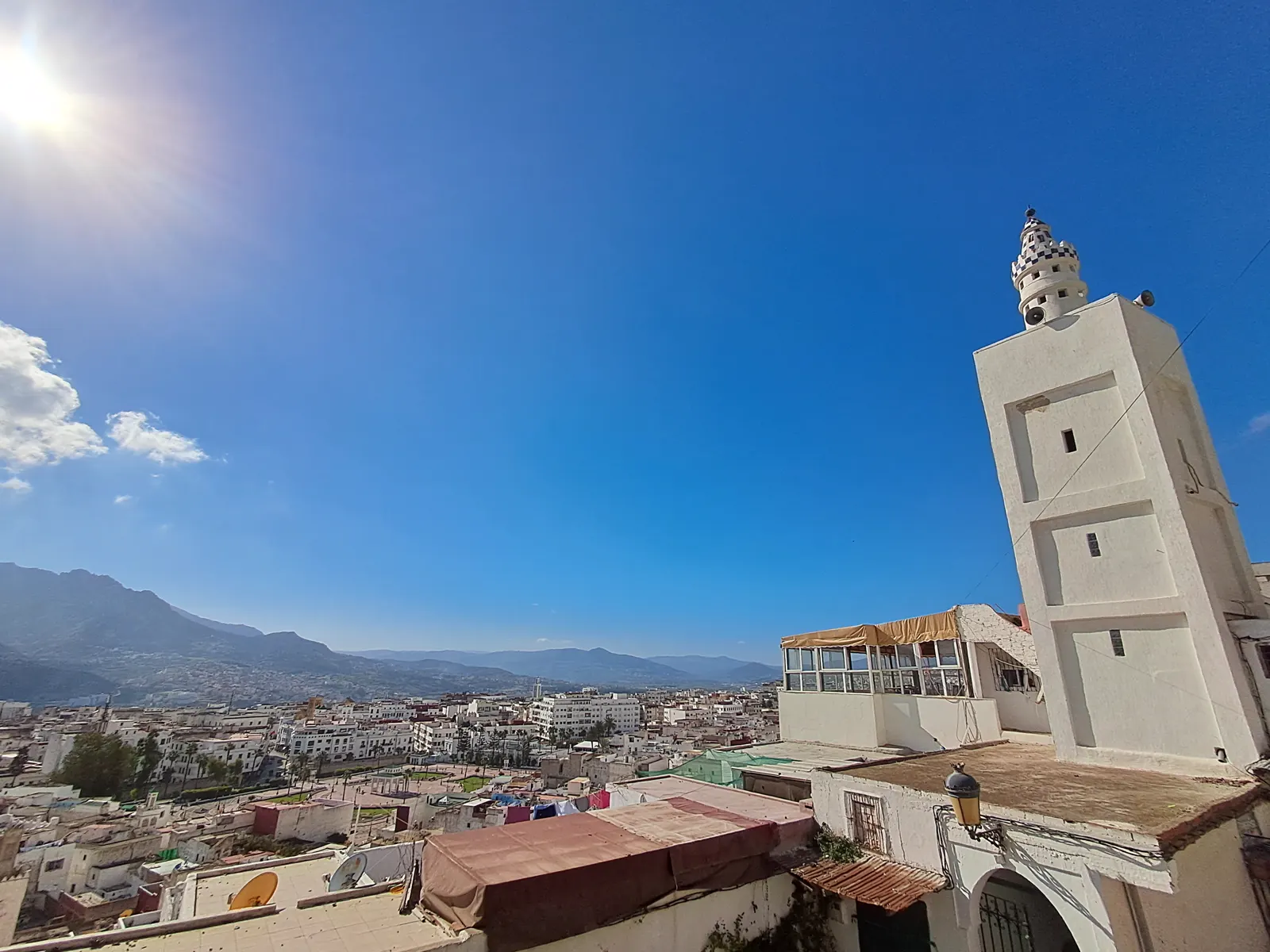 Vista de la ciudad marroquí de Tetuán desde la medina antigua: en primer plano el minarete blanco de una mezquita y azoteas con antenas, al fondo los edificios blancos de la ciudad nueva y las montañas que la rodean bajo un cielo despejado.